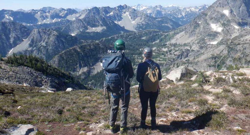 two people wearing helmets and backpack look out over a vast mountain landscape